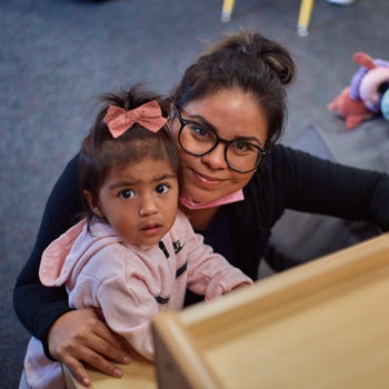 A toddler girl and her mother smile at the camera while playing in a preschool classroom.