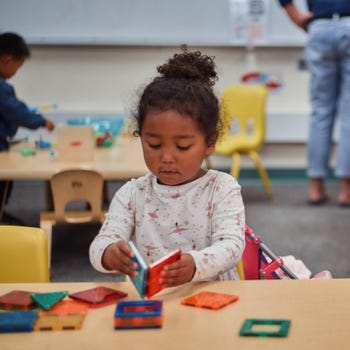 A little girl builds with Magna-Tiles at a table in a preschool classroom.